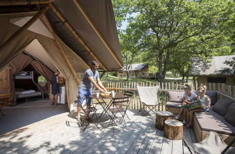 Famiglia si rilassa in una tenda safari Trappeur con terrazza di legno immersa nella natura del villaggio.