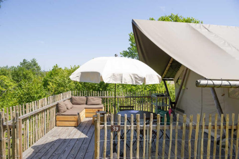 Terrasse mit Sofa, Sonnenschirm und Safarizelt im Village Huttopia Senonches, Frankreich, umgeben von Bäumen.
