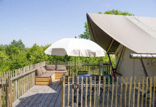 Terrasse extérieure avec canapé, parasol et tente safari au Village Huttopia Senonches, France, entourée d’arbres.