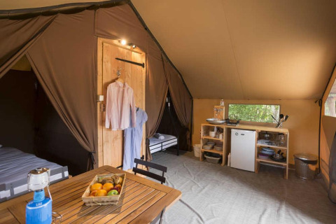 Interior of a luxury safari tent featuring a dining table, fridge, kitchen shelves, and hanging clothes.