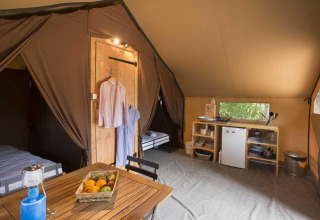 Interieur van een luxueuze safaritent met eettafel, koelkast, keukenrekken en hangende kledij.