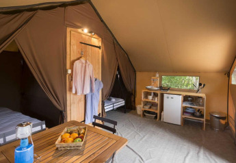 Innenraum eines luxuriösen Safarizelts mit Esstisch, Kühlschrank, Küchenregal und aufgehängter Kleidung.