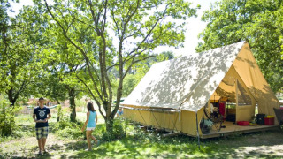 Two people enjoy outdoor camping at a spacious Canadienne safari tent nestled under leafy green trees.