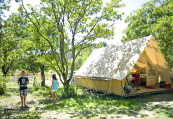 Two people enjoy outdoor camping at a spacious Canadienne safari tent nestled under leafy green trees.