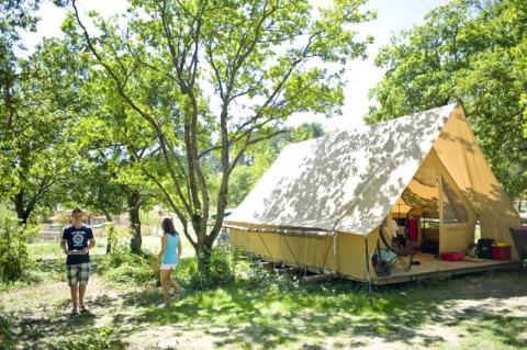Two people enjoy outdoor camping at a spacious Canadienne safari tent nestled under leafy green trees.
