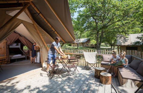 Gezin ontspant op het houten terras van een Tente Trappeur safaritent midden in een groene omgeving.