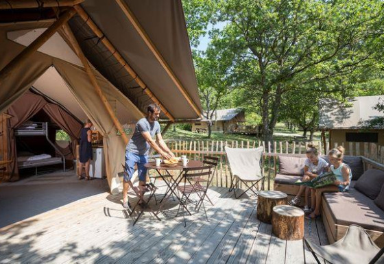 Famiglia si rilassa sulla veranda della tenda safari Tente Trappeur, circondata da alberi verdi.