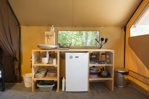 Compact kitchen setup inside a safari tent, featuring a sink, mini fridge, open shelves, and cookware.