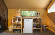 Área de cocina sencilla en una tienda safari, con fregadero, mini nevera, estantes y utensilios.