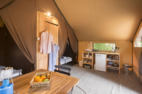 Interno di una tenda safari con tavolo in legno, cesto di frutta, angolo cottura, mini frigo e vestiti appesi.