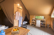 Interior view of a safari tent with a wooden table, fruit basket, kitchenette, mini fridge, and clothes hanging.