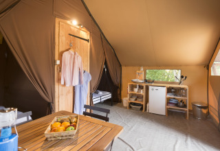 Interior view of a safari tent with a wooden table, fruit basket, kitchenette, mini fridge, and clothes hanging.