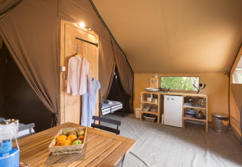 Interno di una tenda safari con tavolo in legno, cesto di frutta, angolo cottura, mini frigo e vestiti appesi.