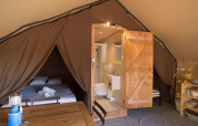 Vista interior de una tienda safari de lujo con cama, baño, mesa de comedor y estante con platos.