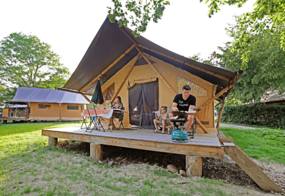 Family relaxing on deck of a Trappeur safari tent, enjoying outdoor dining and grilling together in the sunshine.