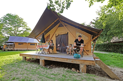 Famiglia si rilassa sulla veranda di una tenda safari Trappeur, grigliando e mangiando all'aperto al sole.