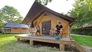 Familie nyder udendørs hygge på terrassen foran en Trappeur safaritelt, omgivet af grøn natur og solskin.