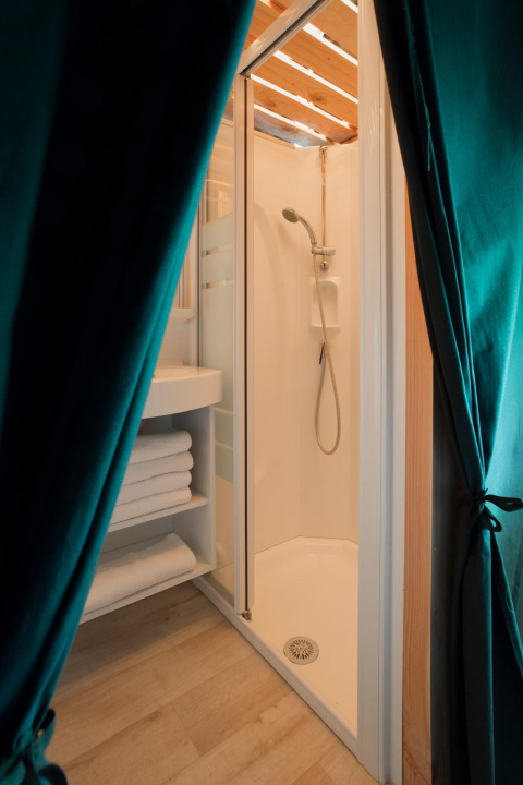 Baño con ducha, estantería de toallas y cortinas verdes en tienda Trappeur de Huttopia Lanmary, Francia.