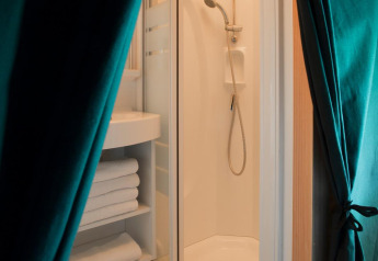 Baño con ducha, estantería de toallas y cortinas verdes en tienda Trappeur de Huttopia Lanmary, Francia.