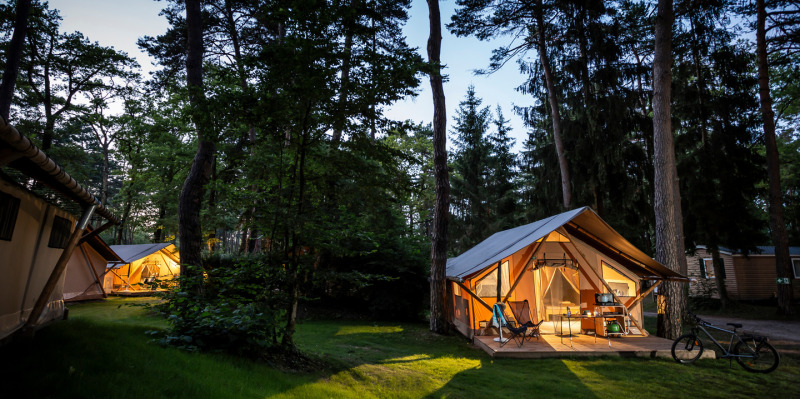 Tentes safari Trappeur éclairées en forêt le soir, entourées d’arbres et d’un vélo à l’avant.