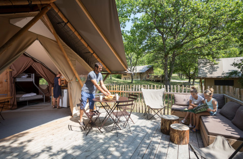 Familia disfruta al aire libre en la terraza de una tienda safari Trappeur, rodeada de muebles y naturaleza.