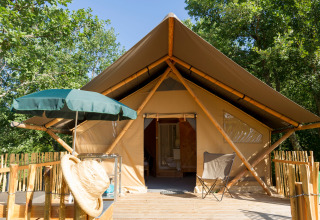 Safari tent with wooden deck, sunshade, hat, and chair in a lush, green forested outdoor setting.