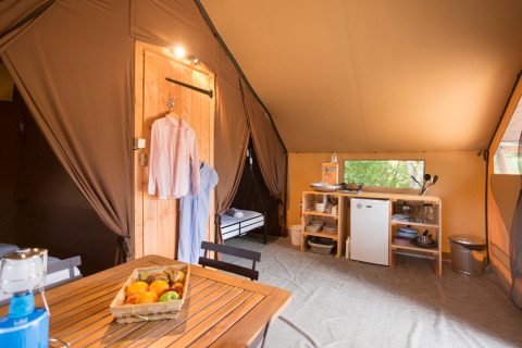 Interior de tienda glamping con mesa de madera, cesta de frutas, cocina pequeña y ropa colgada en la naturaleza.