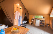 Interior de tienda glamping con mesa de madera, cesta de frutas, cocina pequeña y ropa colgada en la naturaleza.