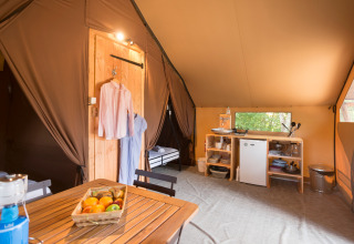 Gezellig glamping-interieur met houten tafel, fruitmand, keukentje en opgehangen kledij in een groene omgeving.