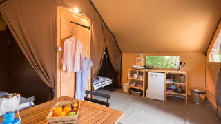 Cozy glamping tent interior with wooden table, fruit basket, kitchenette and hanging clothes in natural setting.