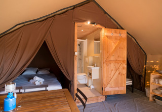 Het interieur van een glampingtent met tweepersoonsbed, badkamer, houten meubelen en keukengerei.
