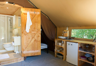 Binnenkant van een safaritent met badkamer, houten deur, kitchenette, kleine frigo en zicht op natuur.