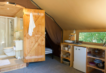 Interno di una tenda safari con bagno, porta in legno, angolo cottura, frigorifero e vista sulla natura.