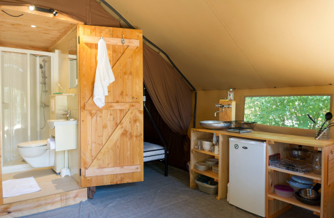 Intérieur d’une tente safari avec salle de bain, porte en bois, kitchenette, frigo et vue sur la nature.