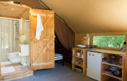 Interior of a safari tent featuring a bathroom, wooden door, kitchenette, small fridge, and nature view.