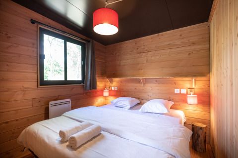 Schlafzimmer mit Holzwänden, Doppelbett, Fenster und roten Lampen im Chalet Toronto, Huttopia Pays de Condrieu, Frankreich.