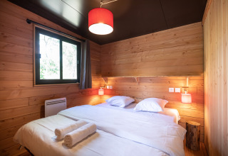 Schlafzimmer mit Holzwänden, Doppelbett, Fenster und roten Lampen im Chalet Toronto, Huttopia Pays de Condrieu, Frankreich.