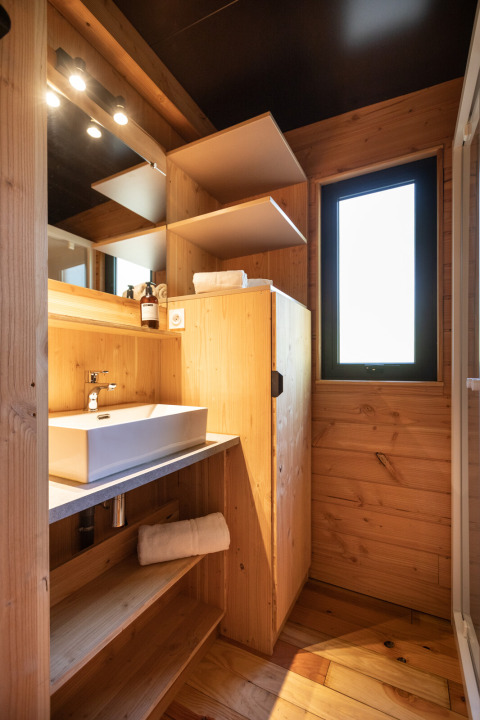 Baño moderno con paredes de madera, lavabo, espejo, estantes abiertos y ventana en Chalet Toronto, Village Huttopia.