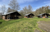 Tres lodges Coco en Camping La Colline, Bélgica, con césped y árboles bajo un cielo azul despejado.