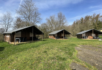 Tres lodges Coco en Camping La Colline, Bélgica, con césped y árboles bajo un cielo azul despejado.