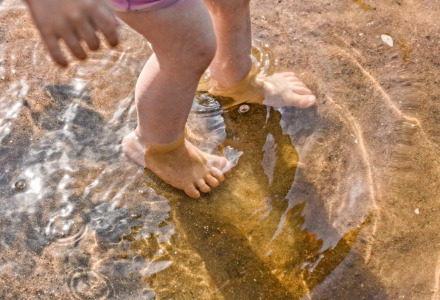 Kinderfüße im flachen Wasser auf sandigem Untergrund in einem Ferienpark mit Glamping-Unterkünften.
