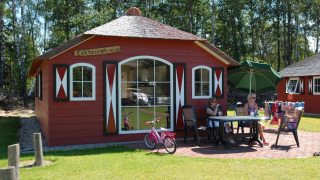 Casa de naturaleza con paredes rojas, personas sentadas afuera en una mesa, bicicleta y paraguas verde.