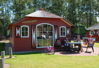Casa nella natura con pareti rosse, persone sedute fuori al tavolo, bicicletta e ombrellone verde.