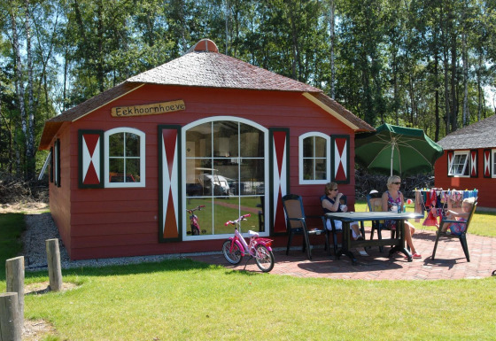 Casa de naturaleza con paredes rojas, personas sentadas afuera en una mesa, bicicleta y paraguas verde.