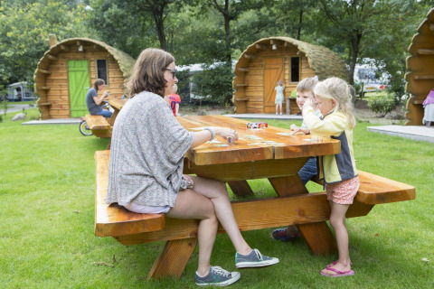 Familie spielt Karten an einem Holztisch vor Baumstammhütten im Veluwecamping De Pampel, Niederlande.