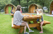 Family playing cards at a wooden picnic table in front of log cabins at Veluwecamping De Pampel, Netherlands.