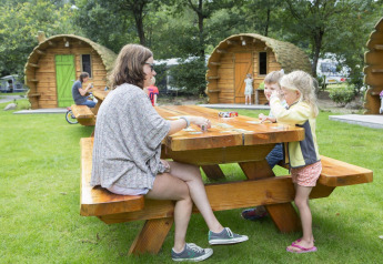 Gezin speelt kaartspel aan een houten picknicktafel voor boomstamhutten op Veluwecamping De Pampel.