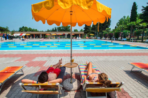 Two people relaxing under a yellow umbrella by the pool at SunLodge Katsura, Camping Gasparina, Italy.