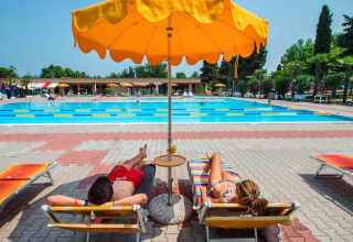 Dos personas descansan bajo una sombrilla amarilla junto a la piscina en SunLodge Katsura, Camping Gasparina, Italia.
