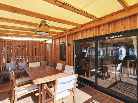 Terrasse couverte d'un lodge avec table à manger en bois, chaises, coussins et portes vitrées coulissantes.